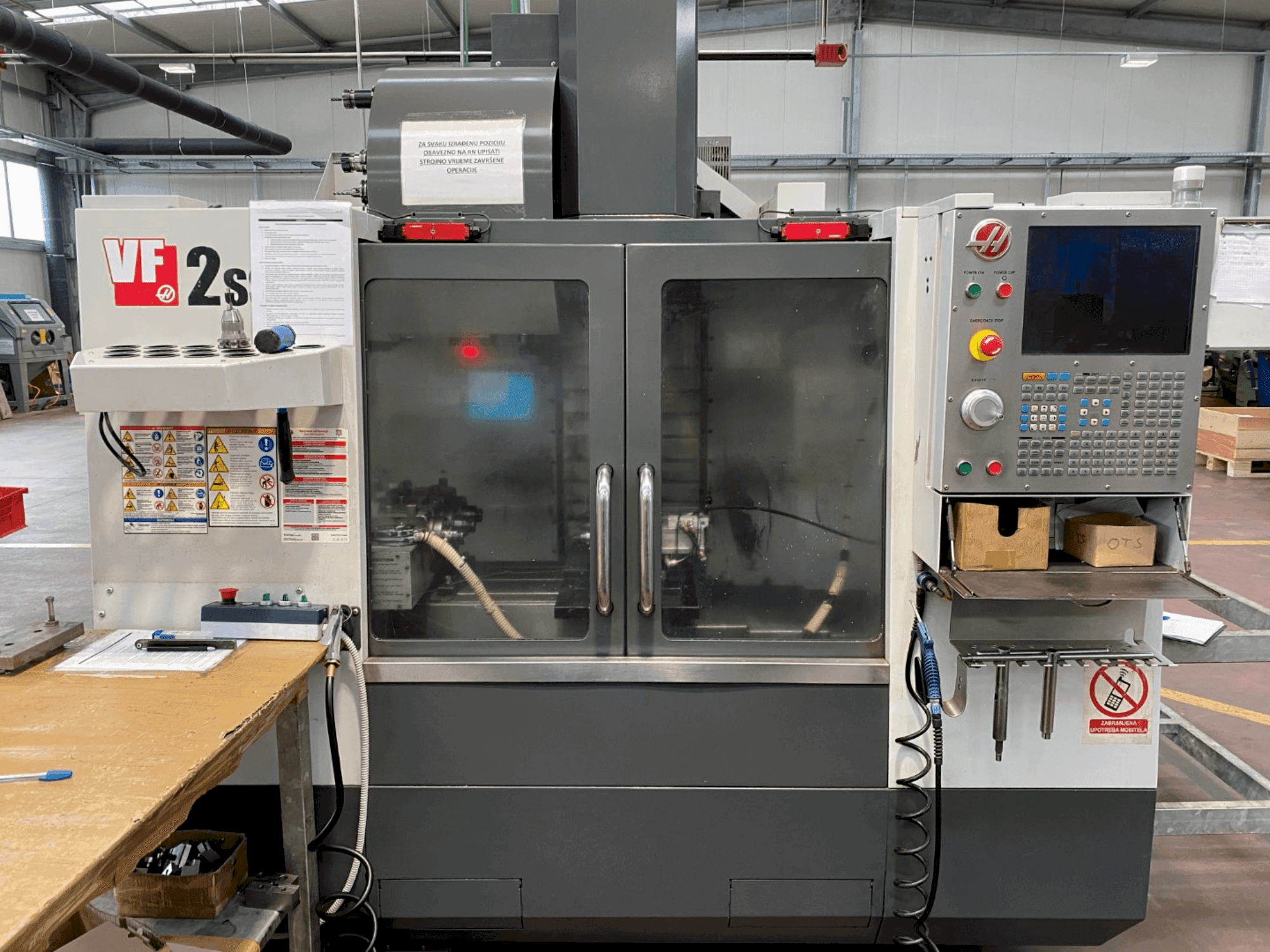 Обрабатывающий центр Haas VF-2SS спереди, панель управления, шланг подачи охлаждающей жидкости и наклейки безопасности сбоку.